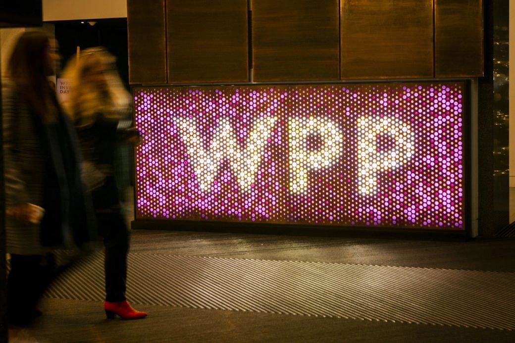 The image depicts a brightly lit display with the letters 'WPP' prominently featured in a colorful arrangement of lights. Two individuals are seen walking past the display, blurred in motion, which adds a dynamic element to the scene.