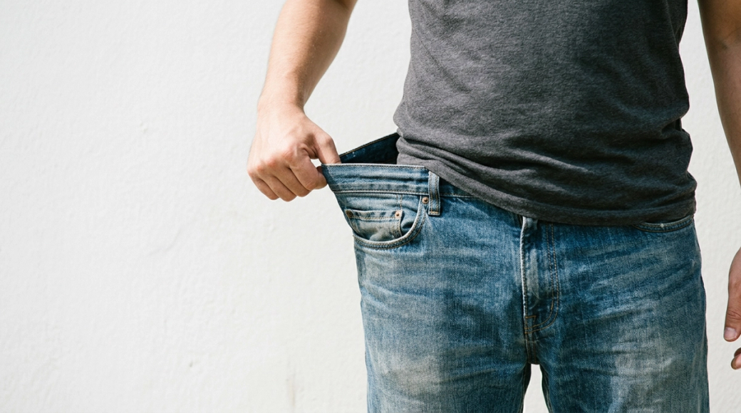 The image depicts a person holding out the waistband of their jeans away from their stomach, revealing a noticeable gap between the fabric and their body. The focus is on the jeans, which appear well-worn, and the casual gesture suggests a lighthearted acknowledgment of significant weight loss. The background is a plain white wall, emphasizing the subject.