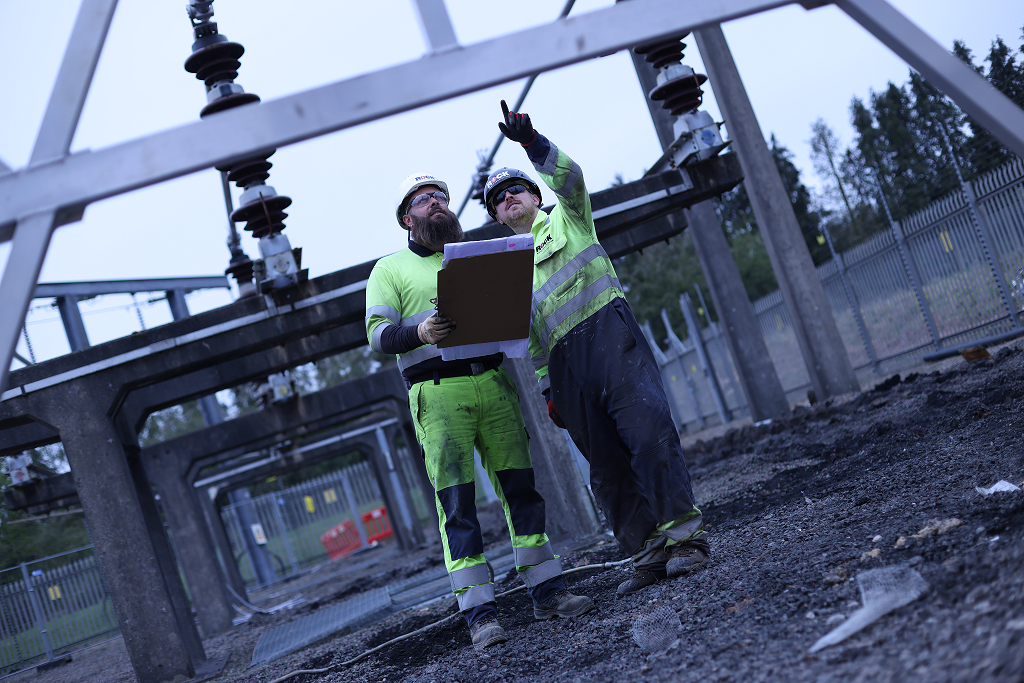 Two workers in high-visibility clothing are inspecting electrical infrastructure at a construction site. One worker is holding a clipboard and pointing at an overhead structure while the other listens attentively, indicating a collaborative assessment.