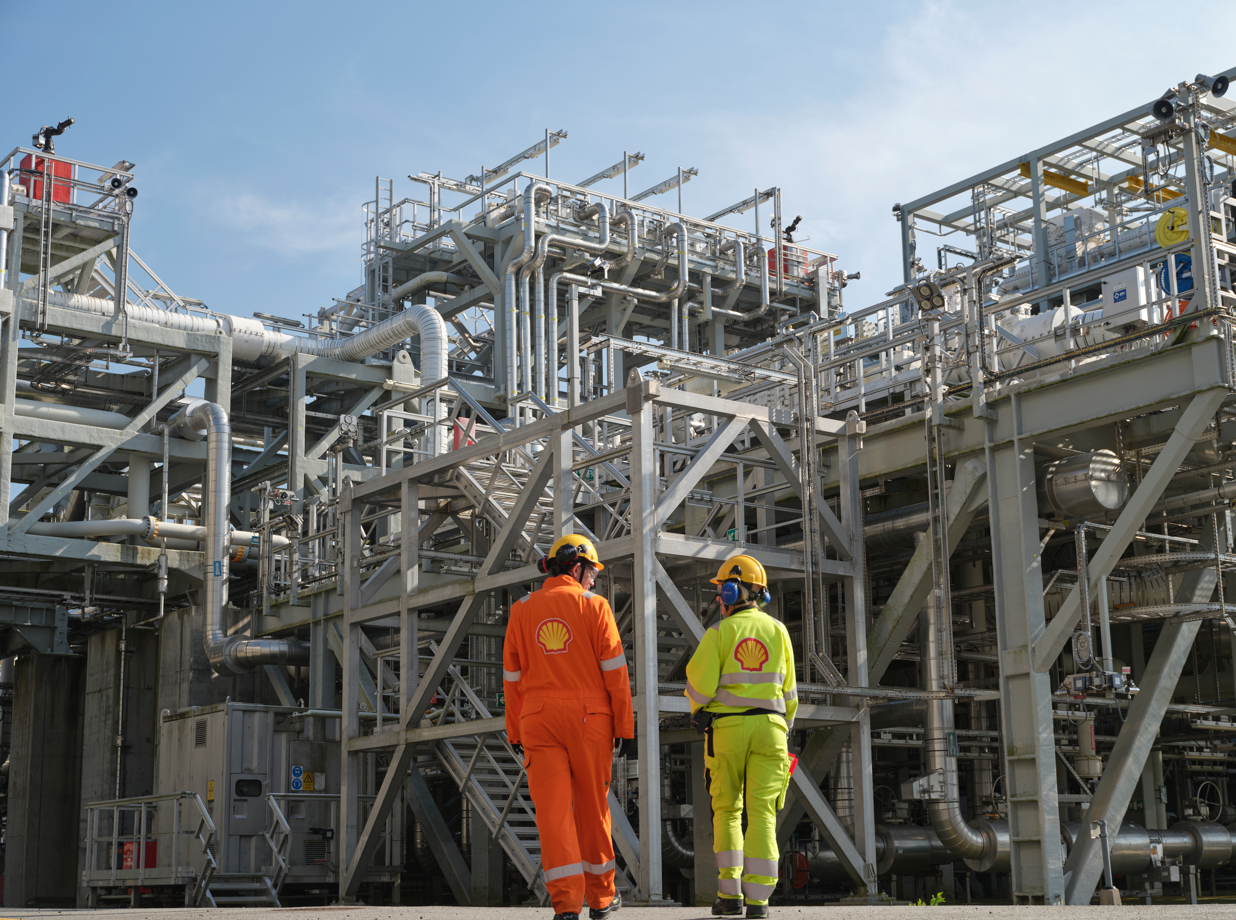 Two workers in protective gear walk through a large industrial facility filled with pipes and machinery. The facility appears to be involved in energy production or processing.