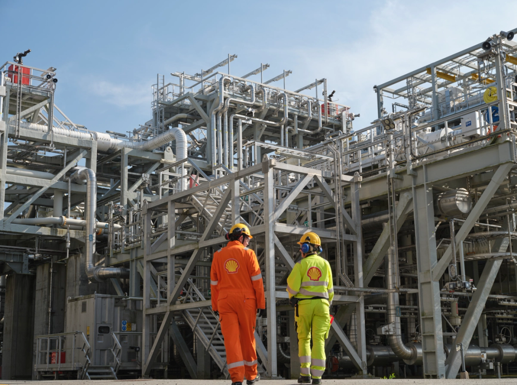 Two workers in protective gear walk through a large industrial facility filled with pipes and machinery. The facility appears to be involved in energy production or processing.