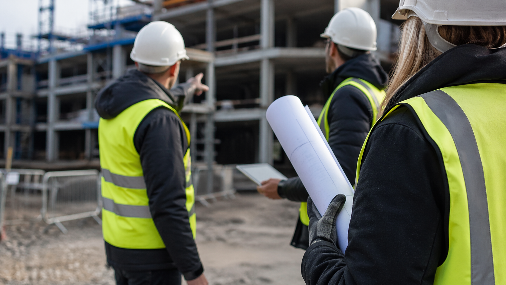 The image shows a group of construction workers in reflective vests and hard hats, observing a building site. One worker points towards the construction, while another holds a set of blueprints and a tablet.