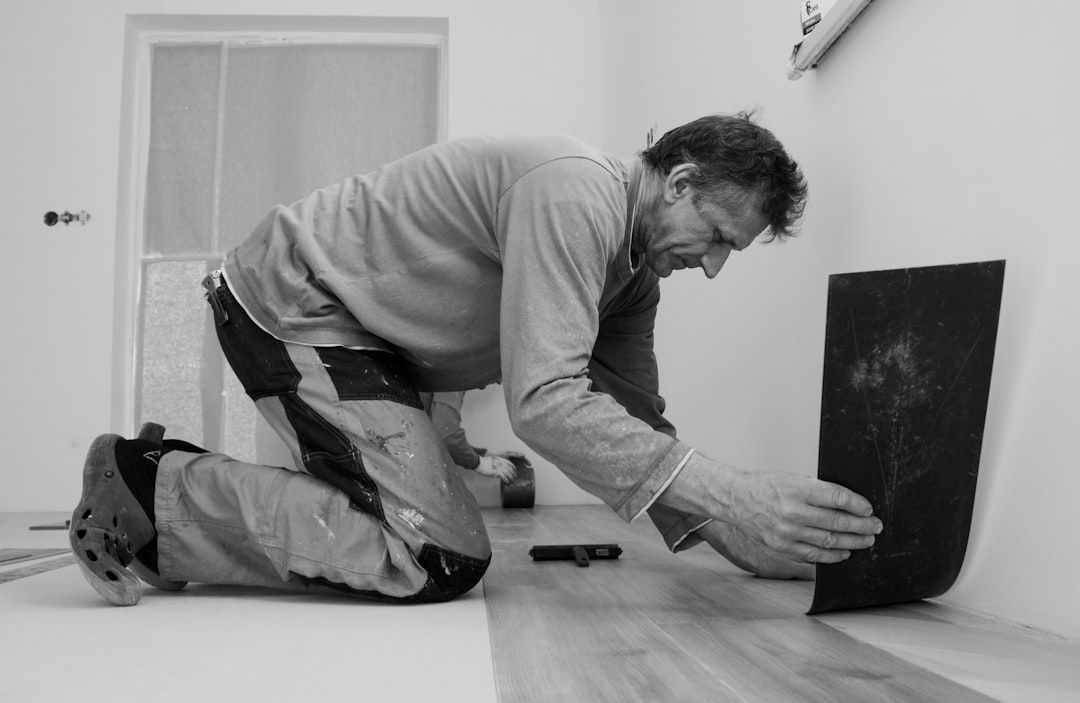 A man kneels on the floor while laying down flooring materials in a room. The image captures the meticulous work of home improvement, highlighting the craftsmanship involved in installing new flooring.