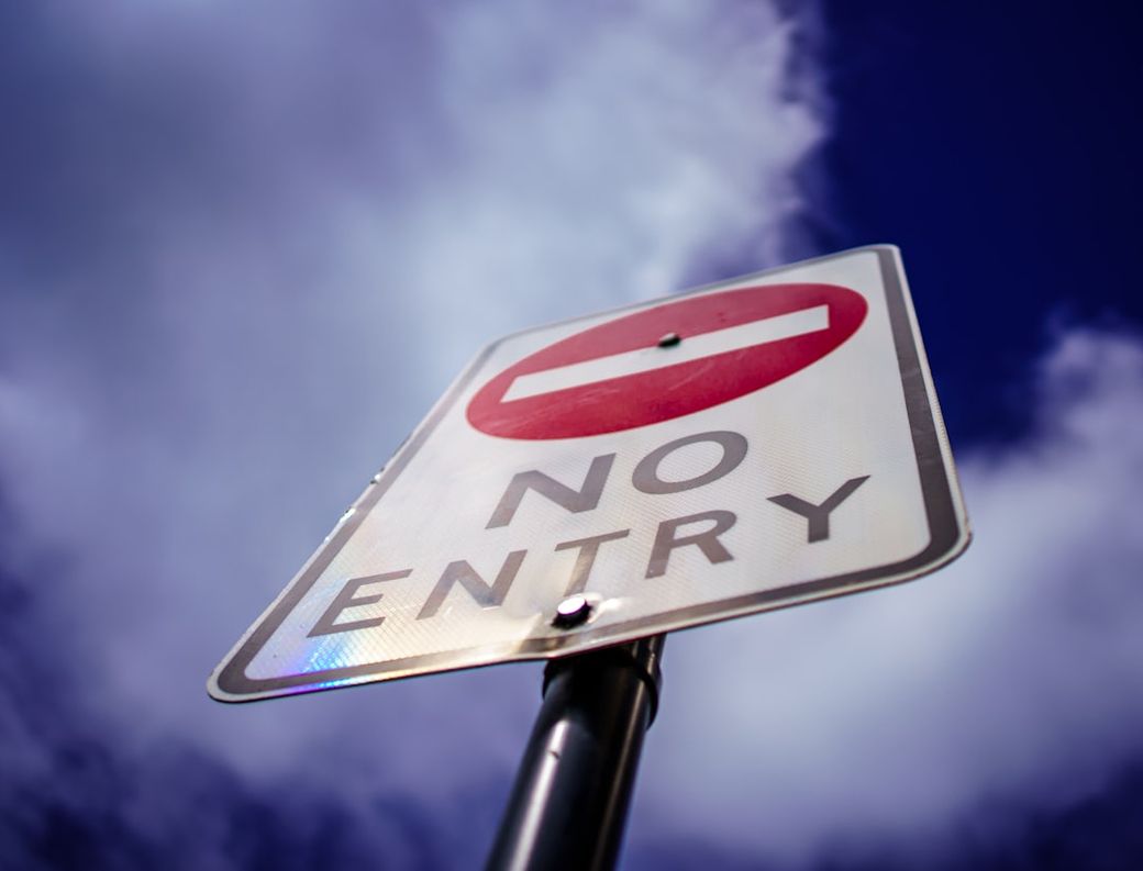 A close-up view of a 'No Entry' road sign against a cloudy sky. The sign features a red circle with a white horizontal bar and clear text indicating restricted access. — Credit: Photo by Joe Pee on Unsplash