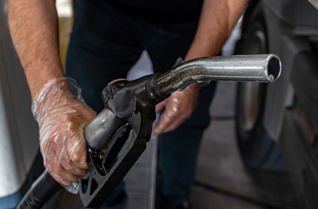 A person wearing disposable gloves is holding a fuel pump nozzle at a gas station. The background shows part of a vehicle, emphasizing the focus on fuel dispensing. — Credit: Photo by Marek Studzinski on Unsplash