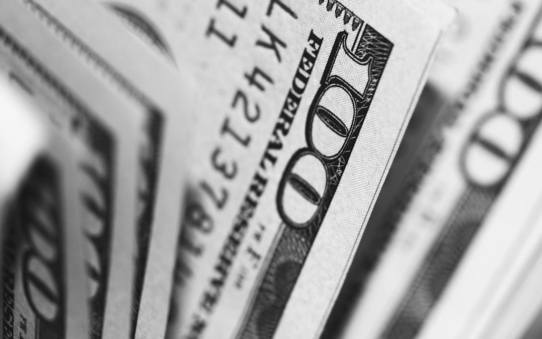 A close-up image of several hundred-dollar bills fanned out, showing intricate details of the currency. The focus highlights the texture and design features typical of U.S. banknotes.