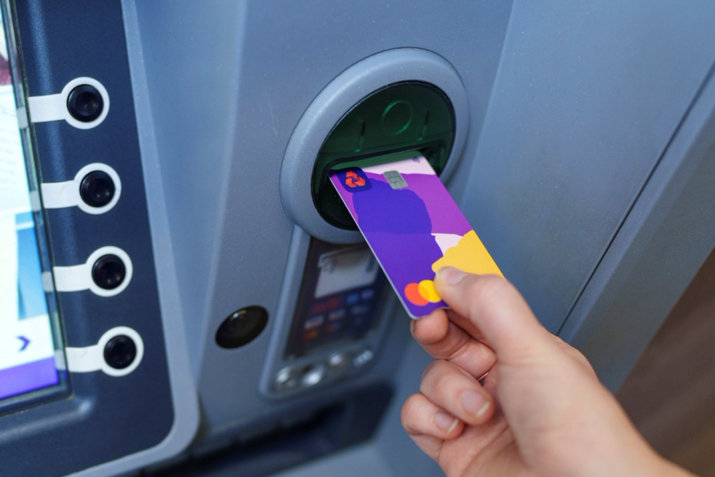 A person is using a colorful card at an ATM to withdraw cash. The ATM machine is modern with a digital display that includes various user interface options. — Credit: Dominic Lipinski/NatWest Group