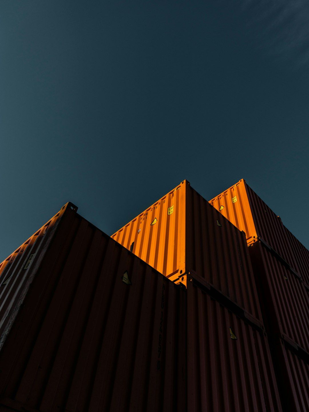 The image depicts a close-up view of stacked shipping containers against a clear blue sky. The containers are primarily orange, casting shadows on each other and highlighting their industrial texture.