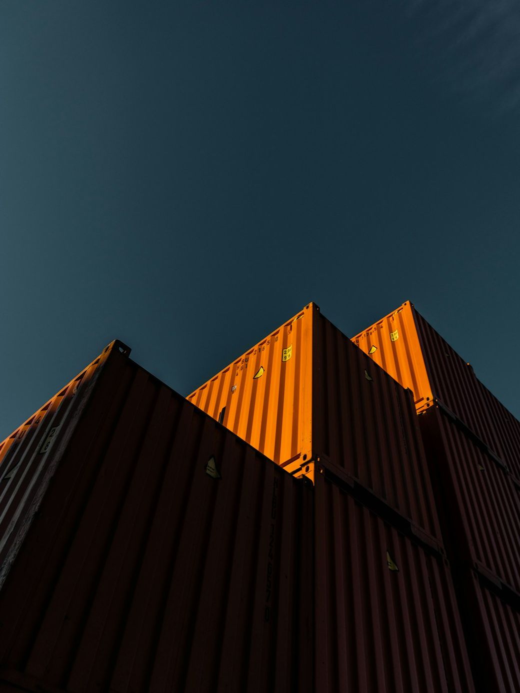 The image depicts a close-up view of stacked shipping containers against a clear blue sky. The containers are primarily orange, casting shadows on each other and highlighting their industrial texture. — Credit: Photo by Aron Yigin on Unsplash