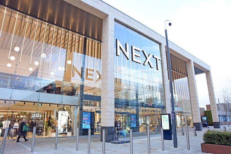 An exterior view of a NEXT retail store, showcasing large glass windows and modern architectural design. The store is situated in a shopping area, with shoppers visible inside and outside.