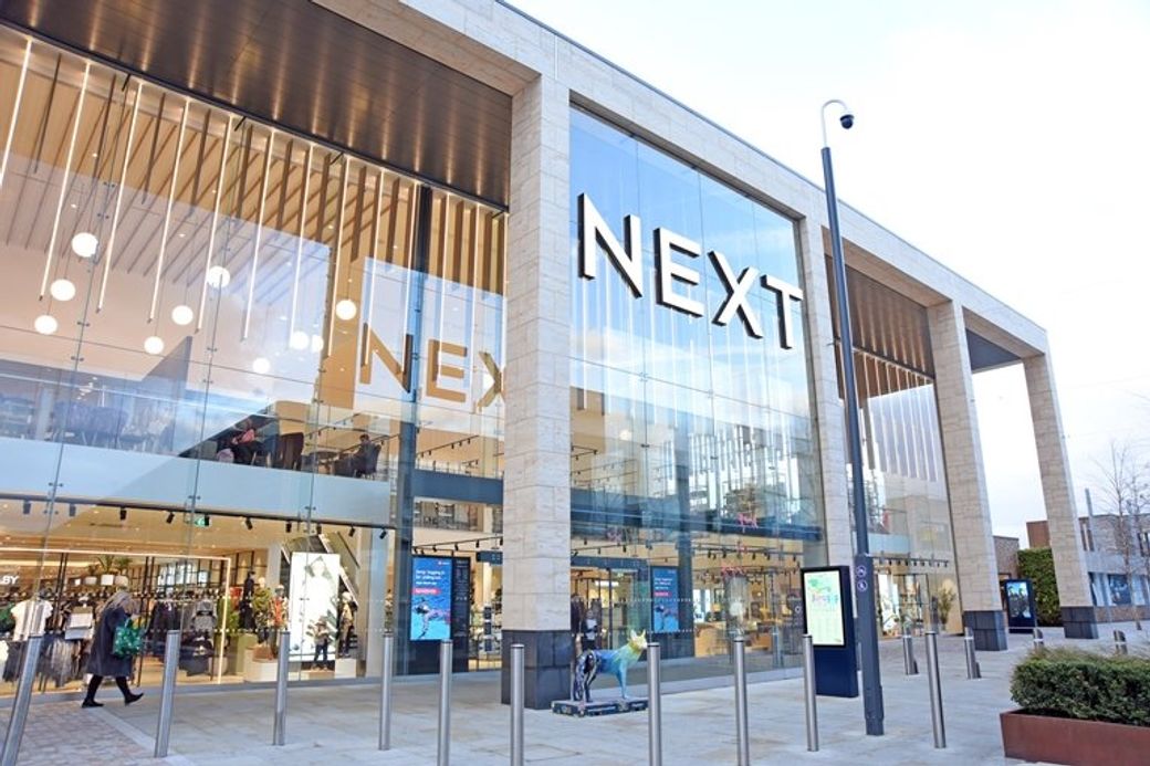 An exterior view of a NEXT retail store, showcasing large glass windows and modern architectural design. The store is situated in a shopping area, with shoppers visible inside and outside.