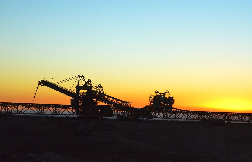 The image captures a silhouette of industrial machinery against a vibrant sunset. The scene depicts a mining site with equipment used for transporting materials in a barren landscape.