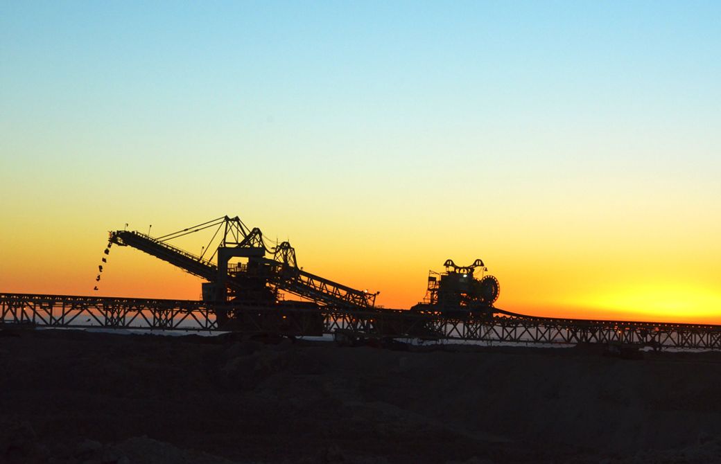 The image captures a silhouette of industrial machinery against a vibrant sunset. The scene depicts a mining site with equipment used for transporting materials in a barren landscape.