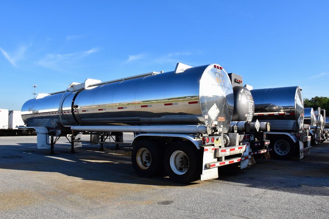 The image depicts a fleet of shiny silver tankers parked in a transportation yard under a clear blue sky. The tankers are designed for transporting liquids, likely fuel or chemicals, and are parked next to detailed tarmac.