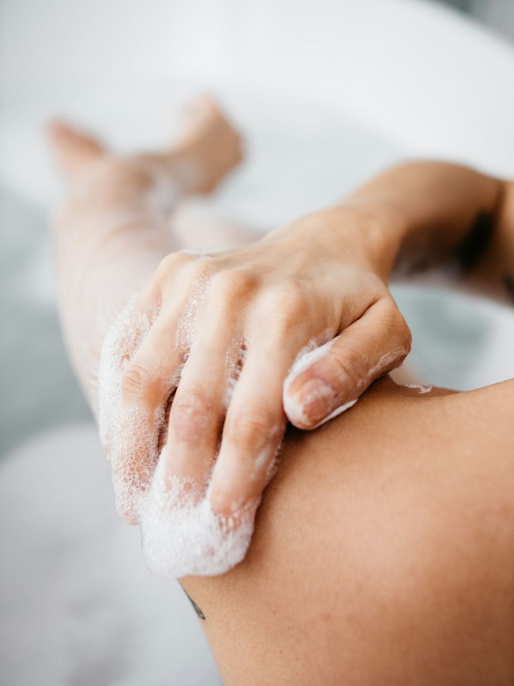The image depicts a close-up of a person's hand applying soap to their skin while in a bath. The focus is on the hand and arm, emphasizing the texture of the soap and water. — Credit: Photo by Redd Francisco on Unsplash