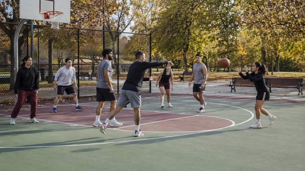 A group of individuals are engaged in a friendly basketball game on an outdoor court surrounded by autumn foliage. Players are actively passing and shooting the ball while enjoying a sunny day.