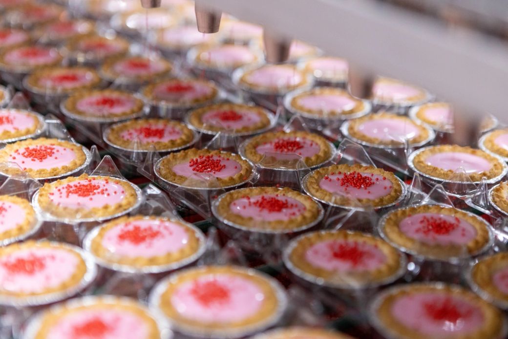 The image depicts a close-up view of a production line filled with small dessert cups. Each cup contains a pink filling topped with red sprinkles, highlighting the process of dessert manufacturing.