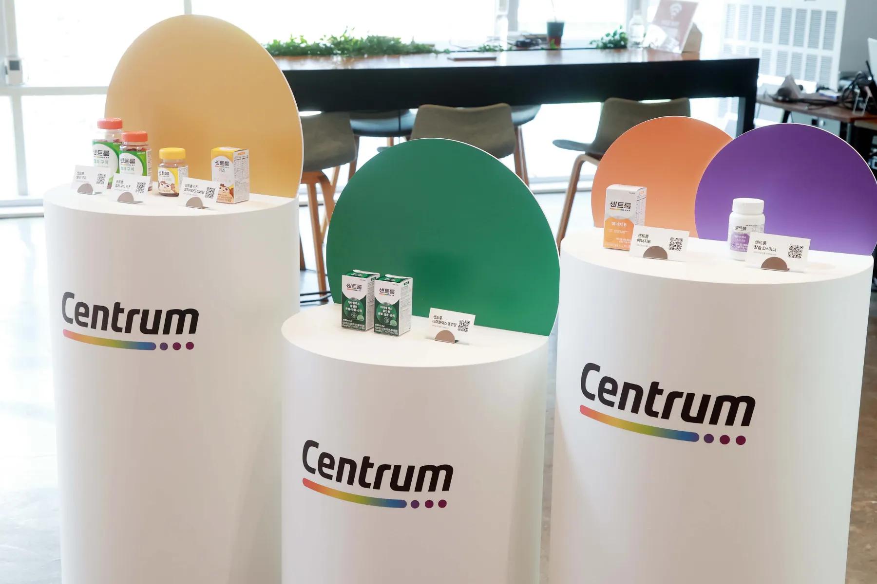 The image shows three cylindrical display stands featuring a range of dietary supplements. Each stand is marked with the brand name 'Centrum' and has products prominently displayed atop.