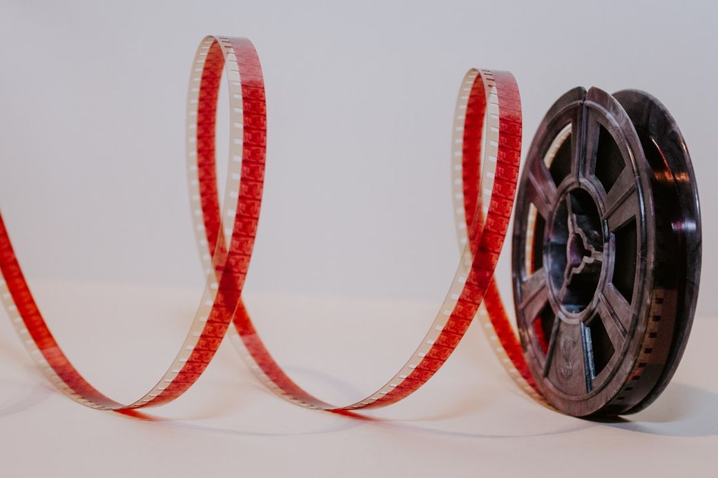 The image features a spool of film with a length of red filmstrip unwound and curled alongside it. The film's vibrant color contrasts with the light background, highlighting the classic medium of movie production. — Credit: Photo by Denise Jans on Unsplash