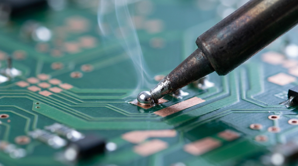 The image features a close-up view of a soldering iron tip making contact with a circuit board joint, where a small bead of molten solder is visible. Smoke rises from the joint as the solder wicks along the copper trace, showcasing the intricate details of electronic assembly.