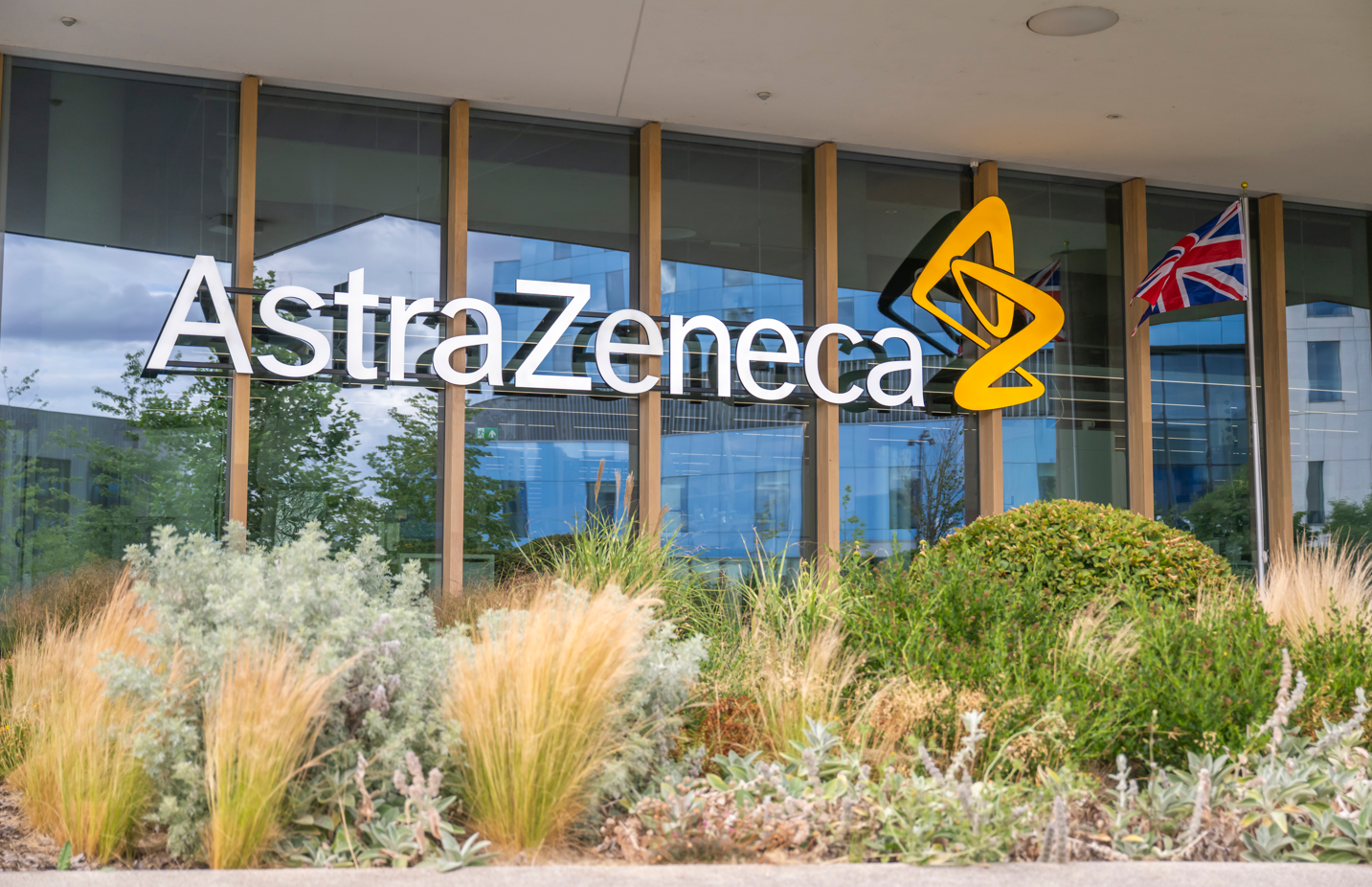 The image depicts the exterior of the AstraZeneca headquarters, showcasing a prominent logo displayed on the building's facade. The area is landscaped with greenery, providing a modern and inviting atmosphere.
