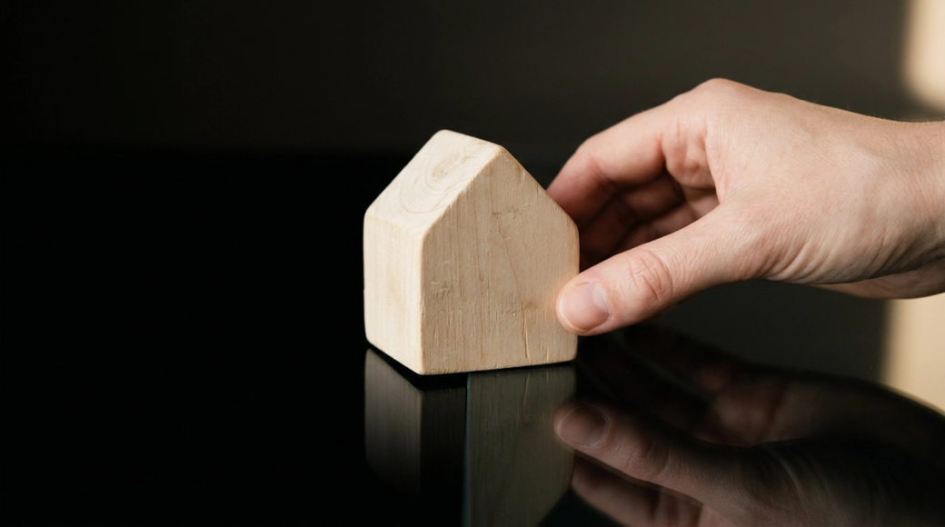 The image features a pale wooden house-shaped block positioned on a dark mirror surface, with a hand sliding the block toward the edge. The sharp reflection of the block contrasts with the matte black void beyond the mirror, suggesting a moment of transition or disposal.