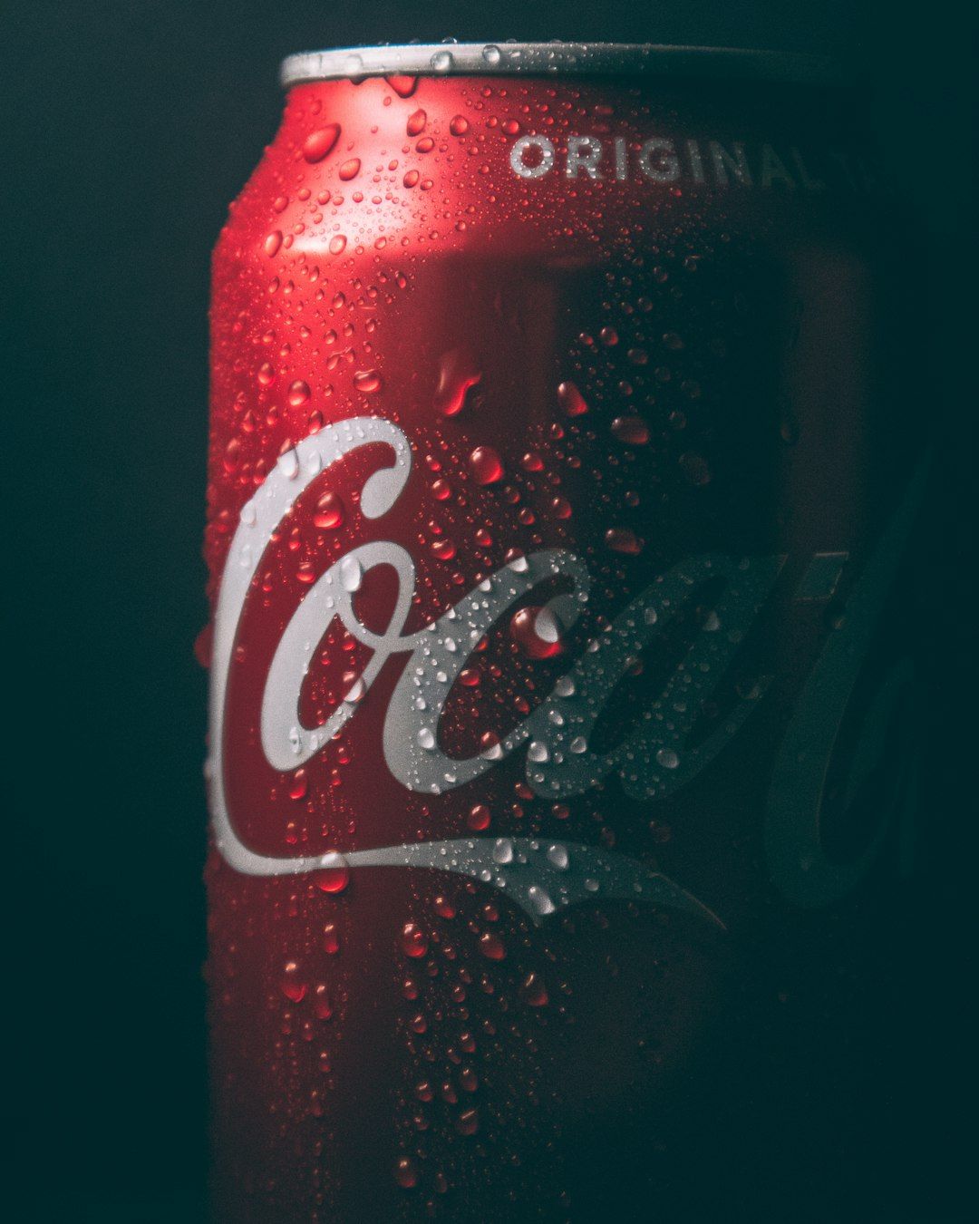 A close-up image of a condensation-covered Coca-Cola can, highlighting its red color and iconic logo. The droplets of water on the surface create a refreshing appearance, emphasizing the drink's cold temperature.