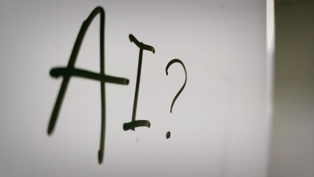 The image features the letters 'AI?' written on a surface, suggesting a contemplation or inquiry about artificial intelligence. The focus is on the text, which appears to be scrawled or drawn in a casual manner. — Credit: Photo by Nahrizul Kadri on Unsplash