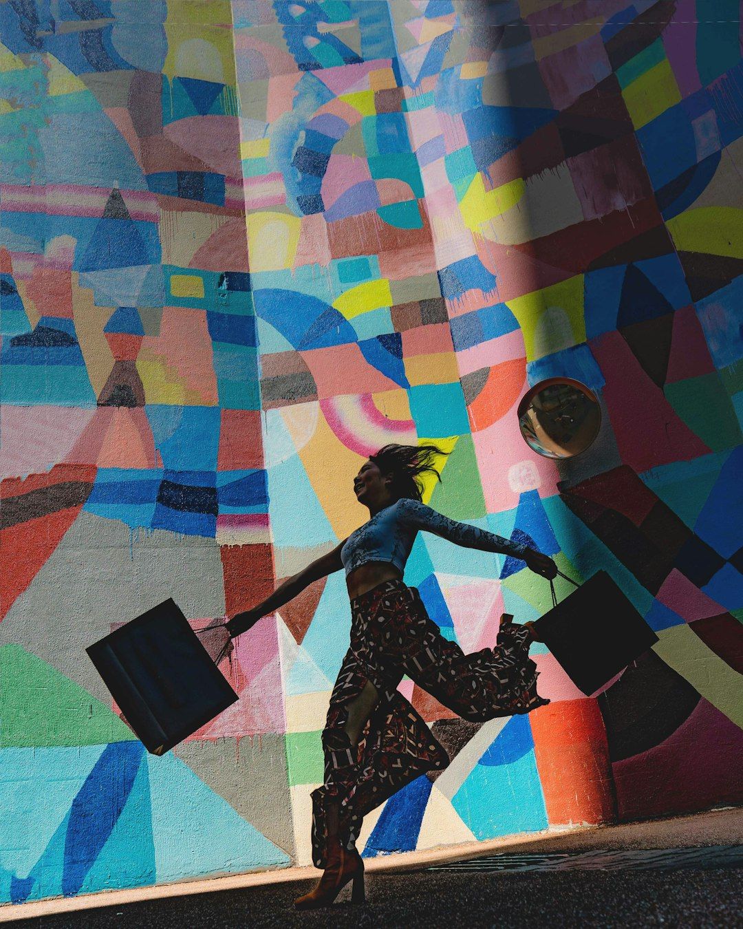 A woman walks energetically against a colorful mural, holding shopping bags in each hand. The vibrant background features an abstract design with bold colors.