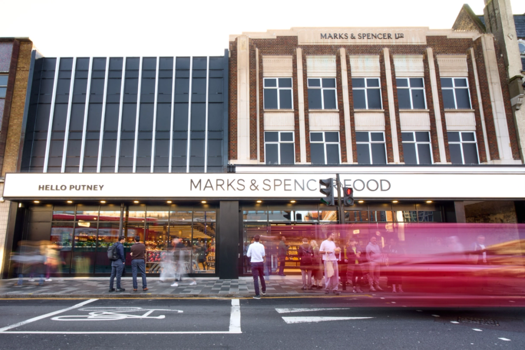 The image showcases the exterior of a Marks & Spencer Food store located in Putney. The scene captures shoppers outside the store, with a red bus passing by, highlighting the bustling urban environment.