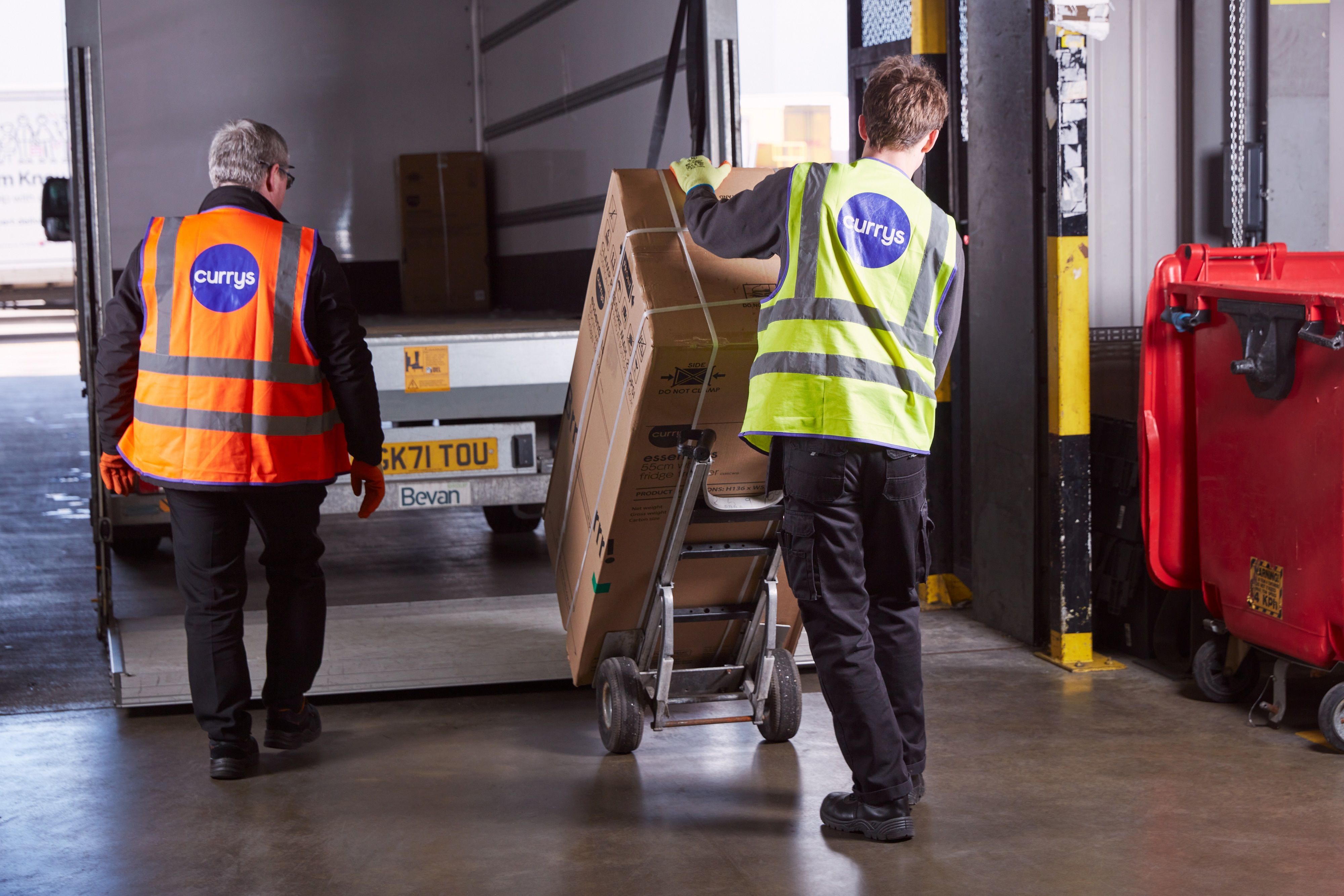 Two workers are seen unloading boxes from a delivery vehicle at a warehouse. One worker is pulling a hand truck with a large box while the other is guiding the process.