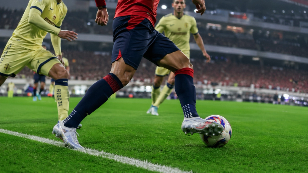 A soccer player prepares to take a corner kick during an intense match, with opponents closely positioned. The vibrant green field and enthusiastic crowd in the background add excitement to the scene.