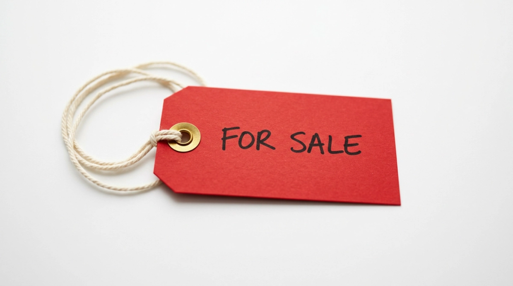 The image features a single red price tag lying flat on a white surface, with 'FOR SALE' hand-written in bold black letters. The brass eyelet and loosely coiled string indicate the tag has been recently detached, embodying a sense of availability and potential transactions in a simplified market context.