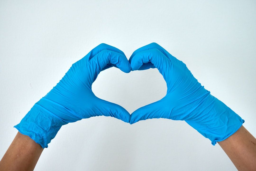 The image features a pair of hands wearing blue latex gloves forming a heart shape against a plain white background. This gesture symbolizes care and compassion, often associated with healthcare and safety.
