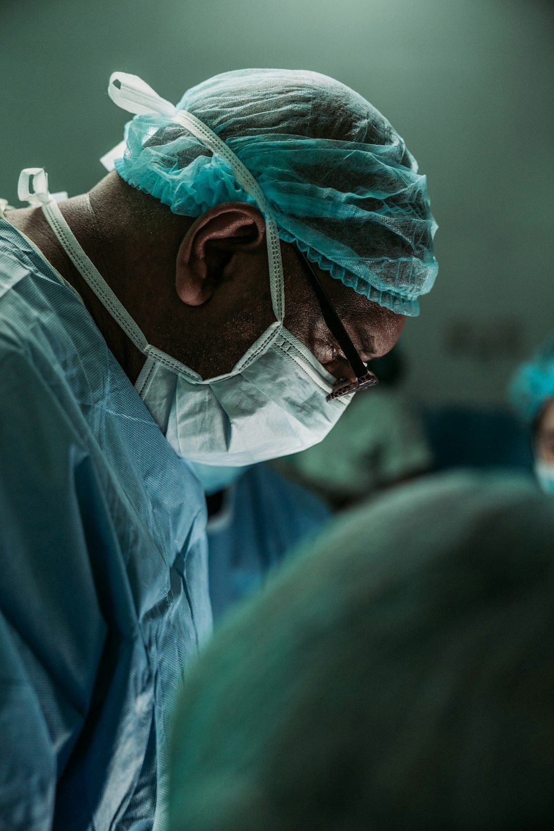 A focused surgeon wearing protective gear, including a mask and surgical cap, is engaged in a medical procedure. The image captures the intensity of a surgical environment, with soft lighting and blurred figures in the background.