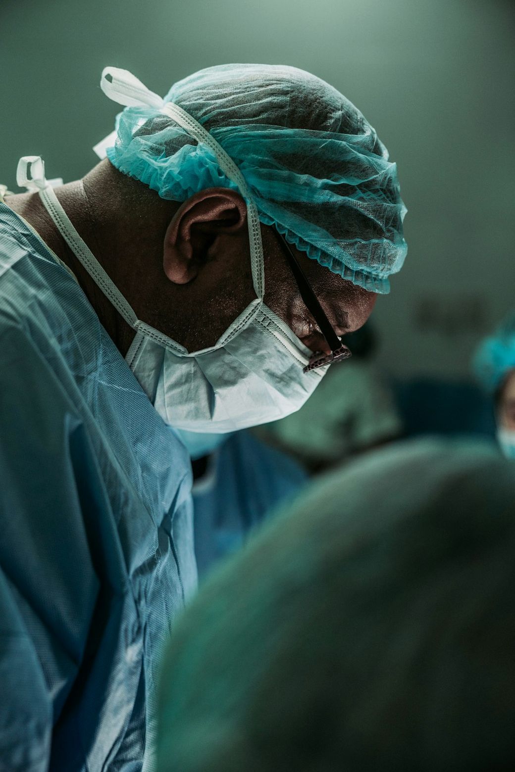 A focused surgeon wearing protective gear, including a mask and surgical cap, is engaged in a medical procedure. The image captures the intensity of a surgical environment, with soft lighting and blurred figures in the background. — Credit: Photo by JC Gellidon on Unsplash