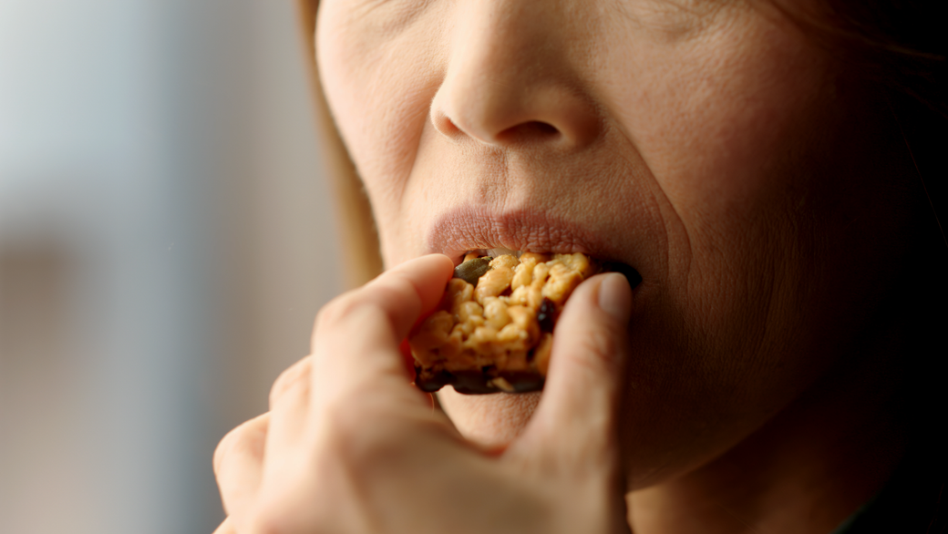 A close-up image of a woman holding a snack bar close to her mouth, preparing to take a bite. The focus is on her lips and the snack, highlighting the act of eating.