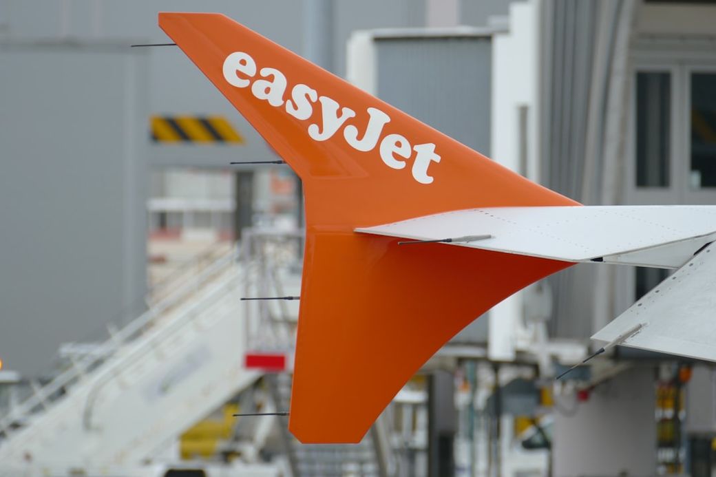 The image features the tail section of an easyJet aircraft, showcasing its distinctive orange and white branding. The background includes elements typical of an airport setting. — Credit: Photo by Call Me Fred on Unsplash