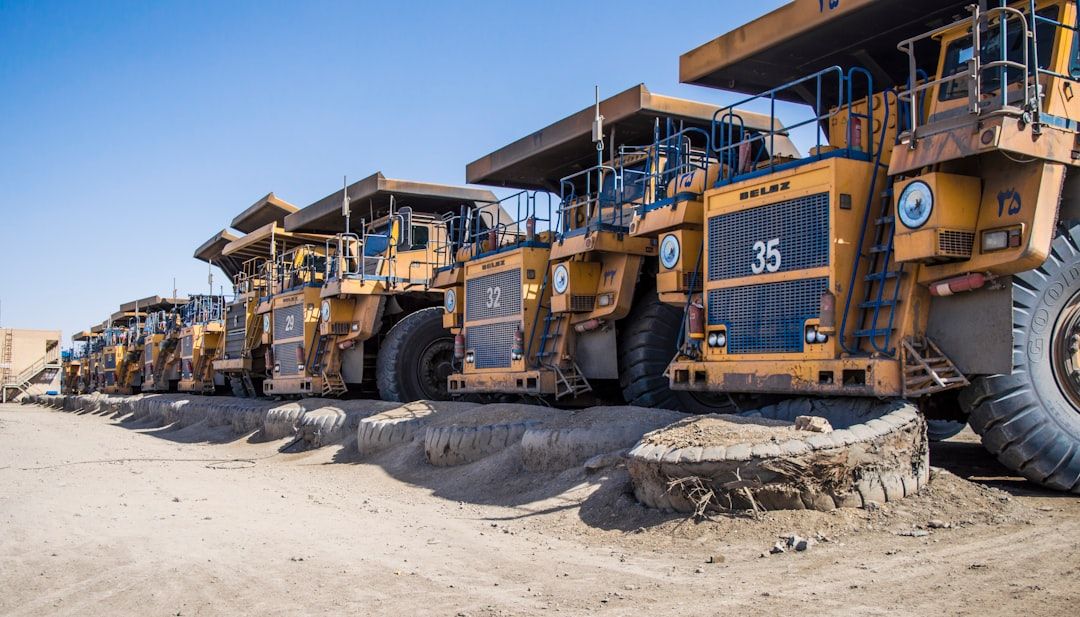 A row of large mining trucks, specifically BelAZ models, parked on a dry, dusty surface under a clear blue sky. The trucks display heavy-duty tires and have a distinct yellow and black color scheme.