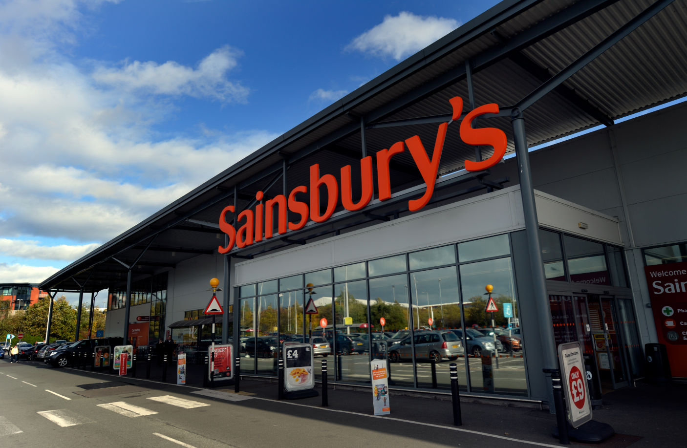 The image shows the exterior of a Sainsbury's supermarket, featuring large signage with the store's name. It is a bright, clear day with clouds in the sky, and the parking lot is visible in front of the building.