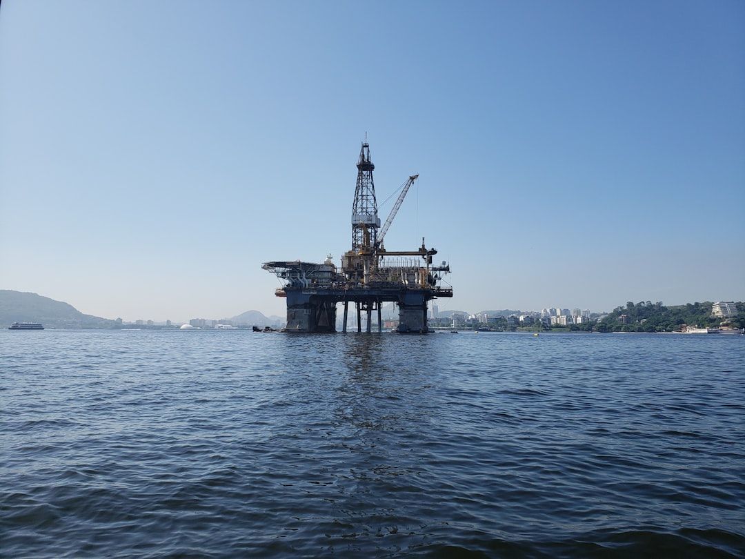 The image shows an offshore oil drilling rig situated in a calm body of water, with a clear blue sky and distant land visible in the background. The rig is depicted in its operational state, highlighting its industrial design.