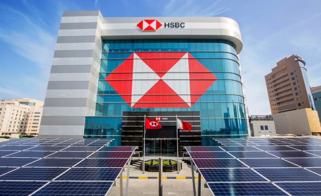 The image shows the exterior of an HSBC bank building prominently featuring the HSBC logo. In the foreground, there are solar panels installed, suggesting a focus on renewable energy.