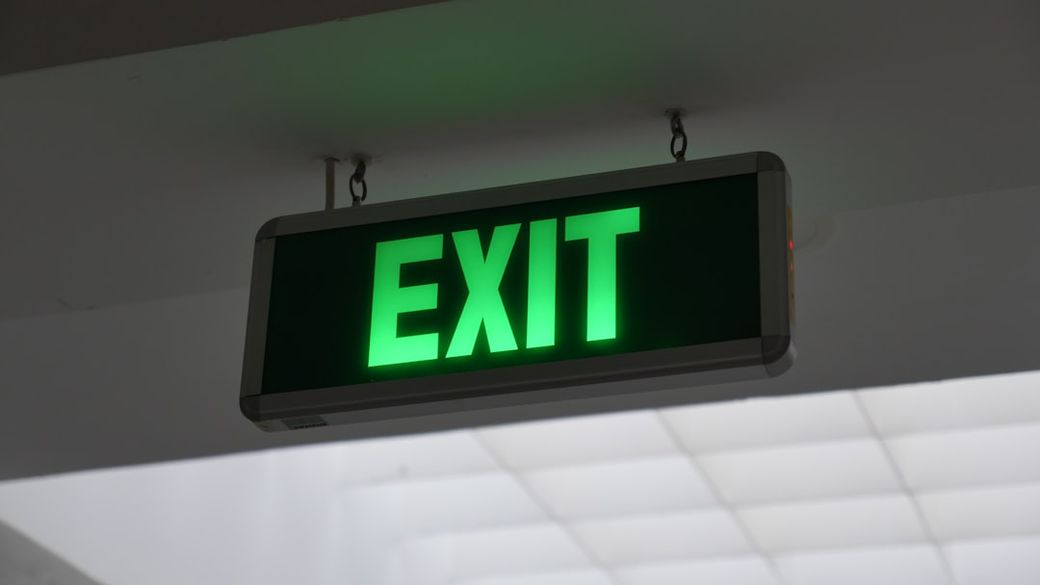 The image features a brightly illuminated green exit sign hanging from the ceiling in a commercial building. The sign is clearly visible against a lighter background, indicating a safe route out. — Credit: Photo by Dongki Koh on Unsplash