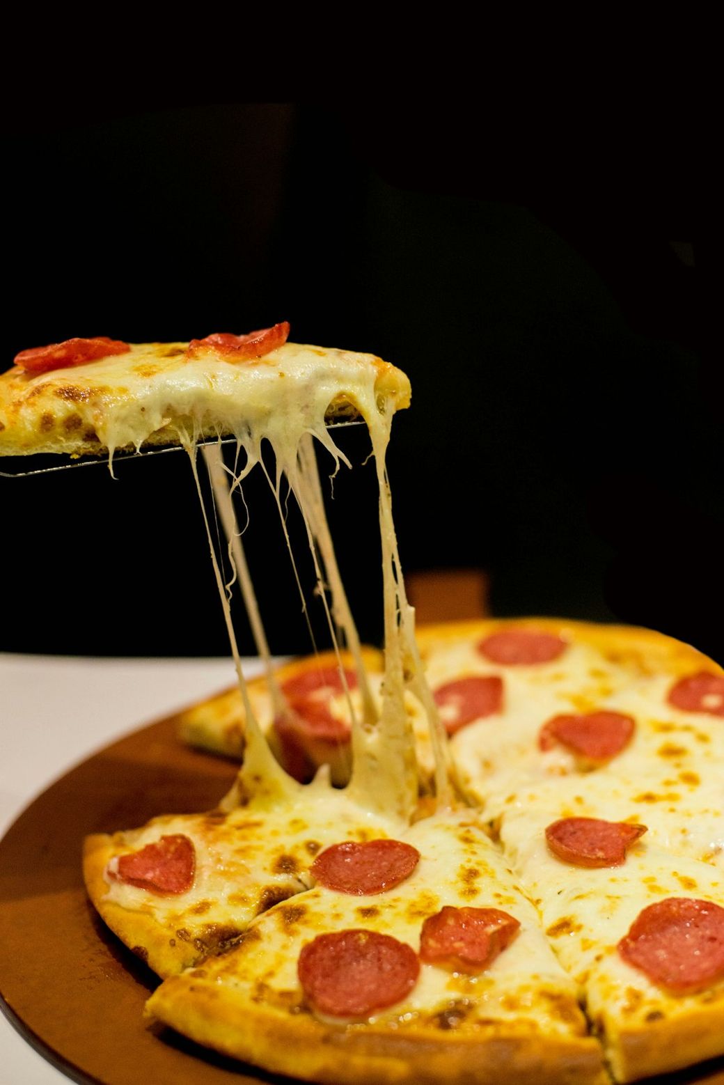 A close-up image of a pizza with pepperoni slices, showcasing melted cheese and a golden crust. The pizza is partially pulled away from the rest of the pie, highlighting the gooey cheese stretch. — Credit: Photo by Fatima Akram on Unsplash