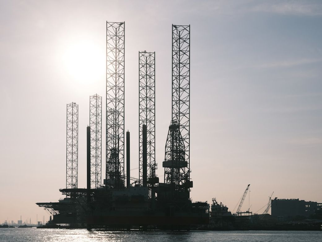 The image depicts an oil drilling rig silhouetted against a sunset, showcasing the structure's towering legs and cranes. In the background, additional industrial elements such as cranes and buildings are visible, emphasizing the maritime industrial environment. — Credit: Photo by Galen Crout on Unsplash