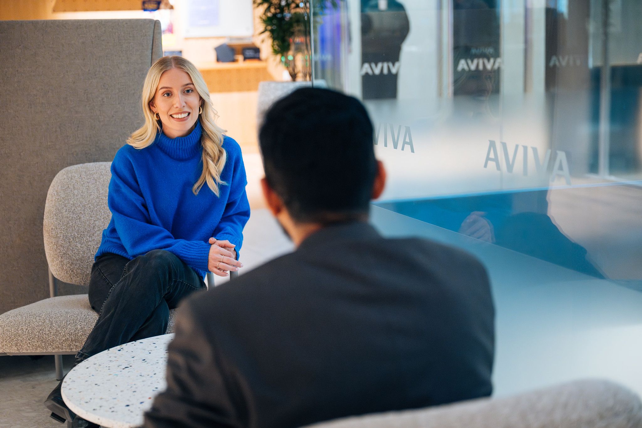 A woman wearing a blue sweater is engaged in conversation with a man, who is sitting with his back to the camera. The setting appears to be a modern office or lounge area, with a glass wall displaying the logo of Aviva.
