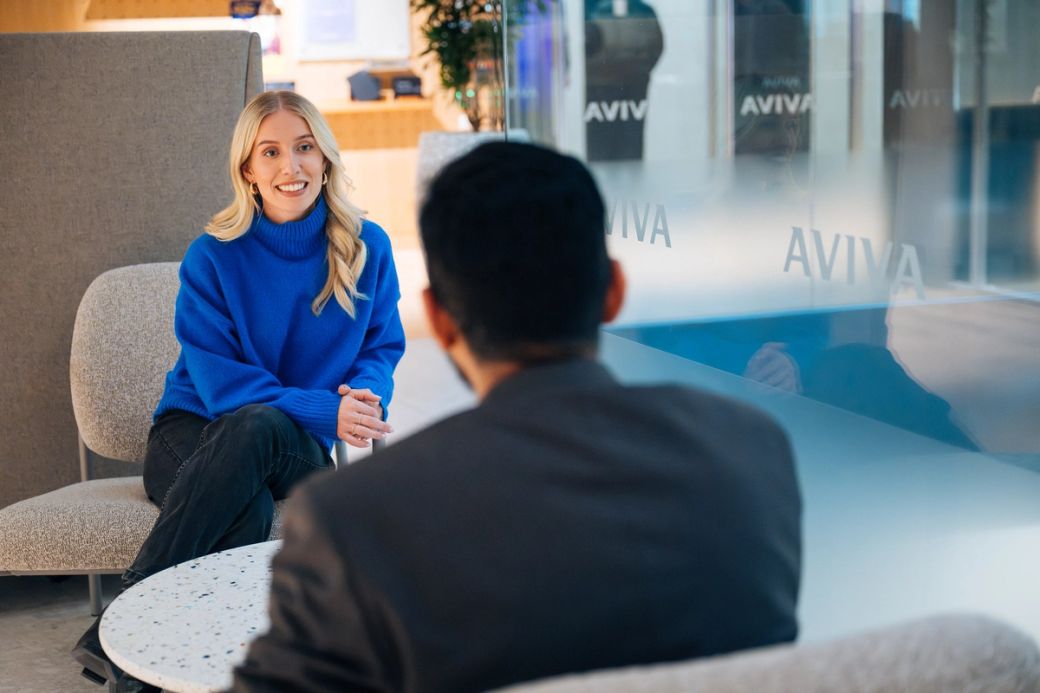 A woman wearing a blue sweater is engaged in conversation with a man, who is sitting with his back to the camera. The setting appears to be a modern office or lounge area, with a glass wall displaying the logo of Aviva.