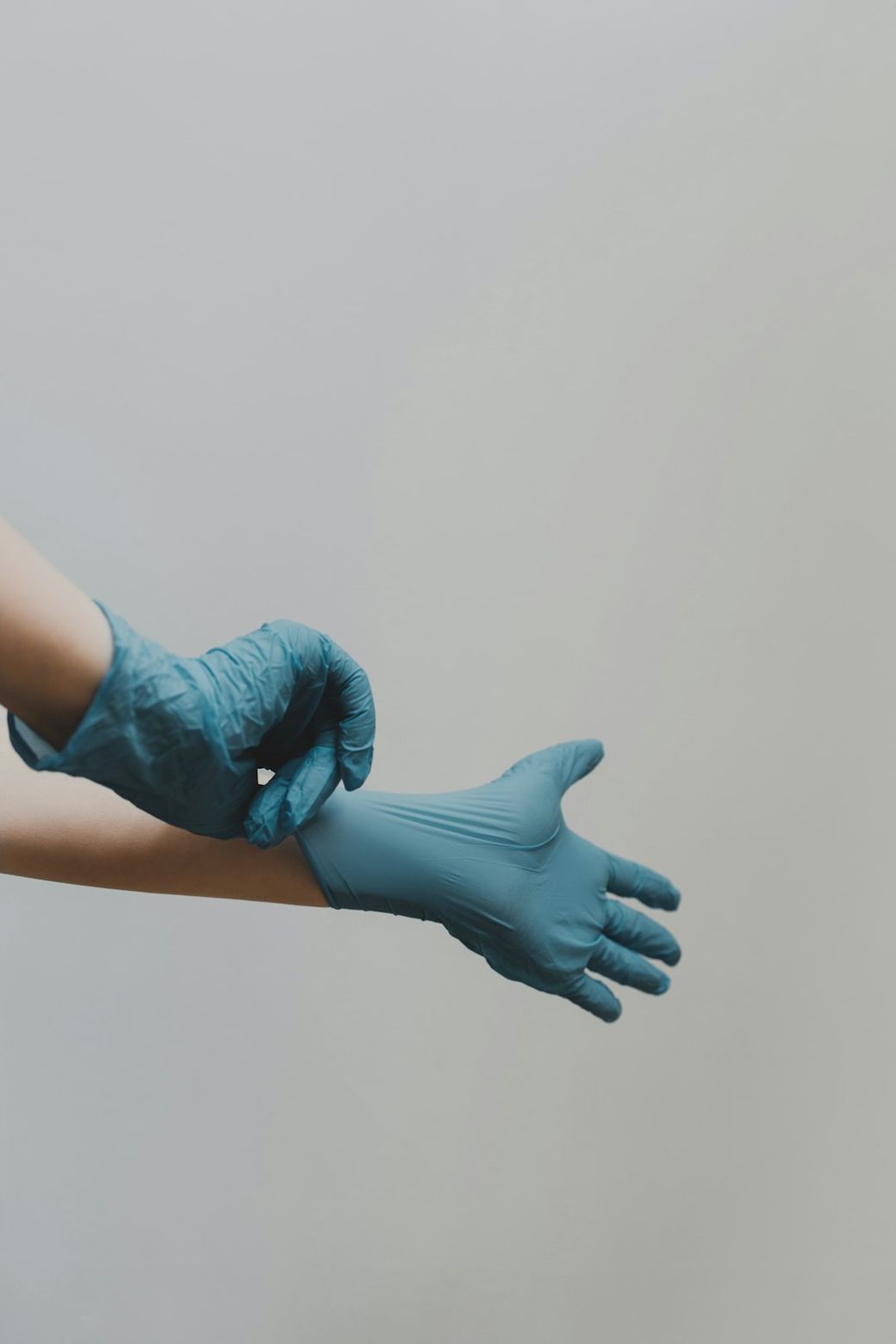 A person is putting on disposable blue gloves, showing the procedure of hand preparation before a medical or hygienic task. The background is neutral and unobtrusive, focusing attention on the hand and gloves. — Credit: Photo by Clay Banks on Unsplash