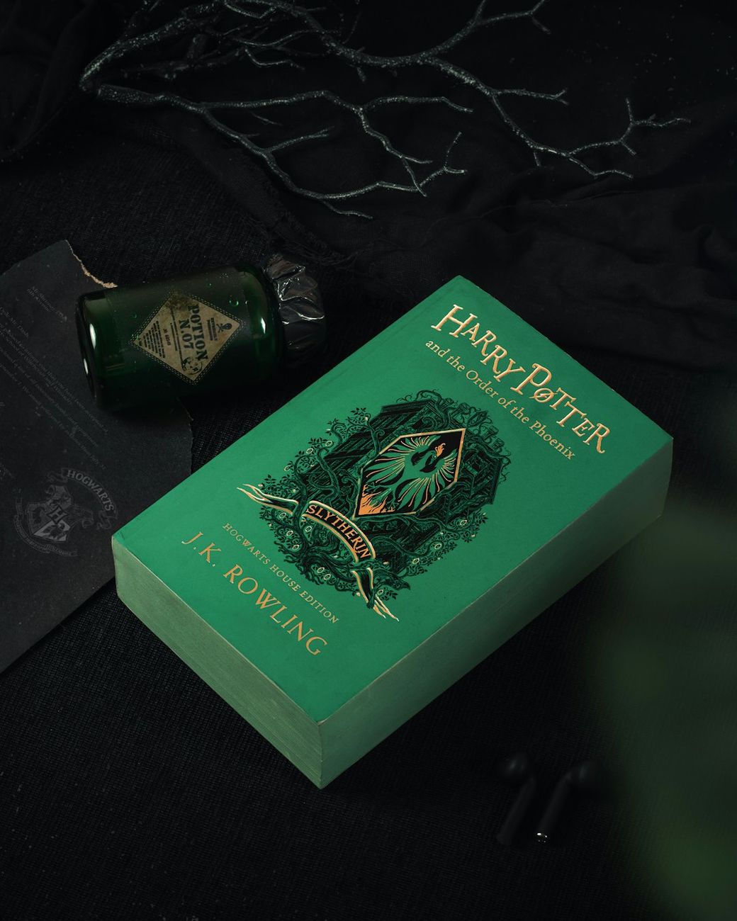 A close-up of a green edition of 'Harry Potter and the Order of the Phoenix' by J.K. Rowling, placed on a dark textured surface. Nearby, there is a vintage-looking bottle, adding a mystical atmosphere to the composition. — Credit: Photo by shri on Unsplash