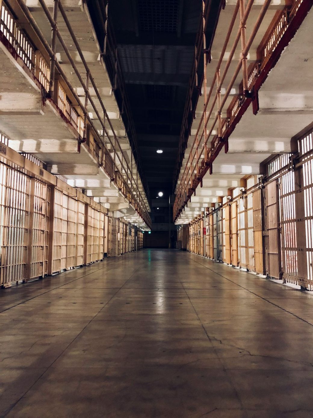 The image features a long, empty corridor within a prison facility, with barred cell doors lining both sides. The lighting creates a dramatic contrast between the shadows and the illuminated areas, emphasizing the stark environment. — Credit: Photo by Carles Rabada on Unsplash